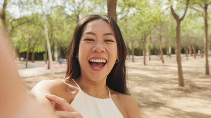 Smiling brunette girl dressed in white top and jeans, taking selfie on mobile phone while standing in city park