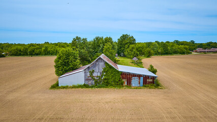 Wooden and metal barn rusting with three empty dirt fields and a patch of green forest behind aerial © Nicholas J. Klein