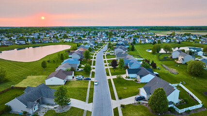 Sunset suburban neighborhood with pond one-story and two-story houses green lawns visible sun aerial