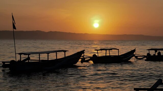 The silhouette of a line of fishing boats moored on the Pangandaran beach being rocked by the waves as the rising sun appears round above the skyline. Slow motion and handheld
