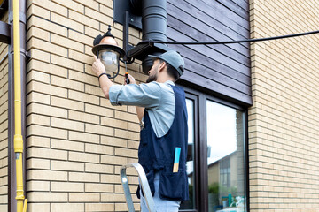 Young handsome electrician standing on ladder and change the light bulb in house facade on the...