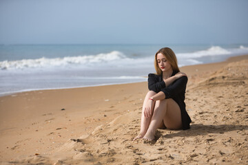 Fototapeta na wymiar Young, blonde, beautiful woman in a bikini and black colored shirt sitting on the beach, sad, lonely, sorry, depressed. Concept loneliness, sadness, grief, depression.