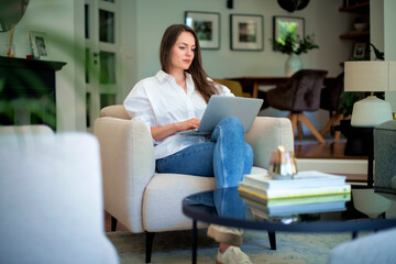 Attractive young woman sitting at home in an armchair and using laptop for work