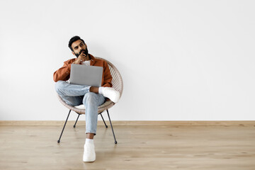 Thoughtful indian man using laptop sitting in armchair indoor