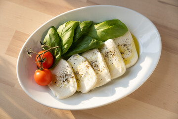 Fresh mozzarella, tomatoes, and sweet Italian basil on a white plate next to a kitchen window at sunset 