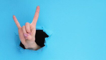 Horns sign. Rock cool. Female fan hand showing fingers up inside breakthrough paper hole on blue ripped wall background with empty space.