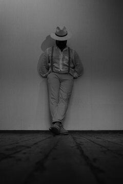 A strict man with a beard in a white suit and white hat stands cross-legged and leans against the wall. The gaze is directed to the floor, hands in pockets. Vertical black and white photo.
