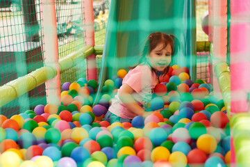 Obraz premium Children's street play attraction fenced with a net. A little girl rolled down the slide and is floundering among many colorful balls.