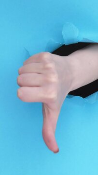 Vertical video. Vote gesture. Dislike sign. Female hand showing rejection no thumb down on blue breakthrough paper hole ripped wall background with copy space loop.