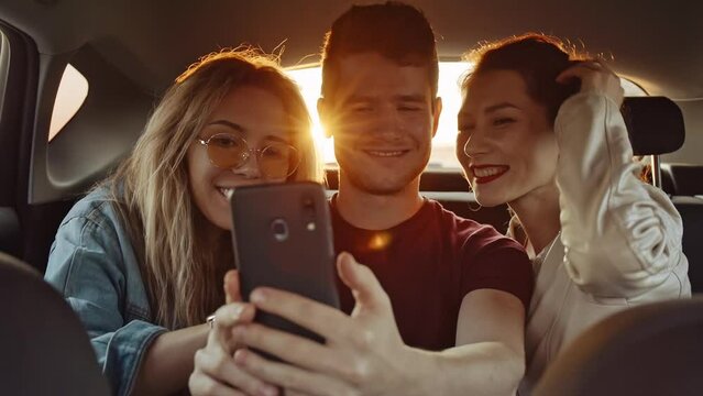 Handsome young guy making selfie with pretty girls on phone or smartphone in back seat of moving car while roadtrip. Hipster blogger taking photo for social network. Concept of social media.