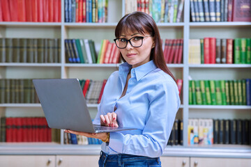 Portrait of confident female teacher holding laptop inside library