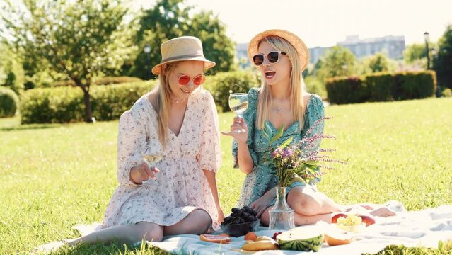 Two young beautiful smiling hipster female in summer sundress and hats. Carefree women making picnic outside. Positive models sitting on plaid on grass, drinking wine, eating fruits. Cheerful, happy