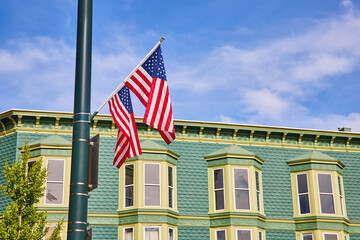 Two American flags on pole with blue sky and a green tree in front of teal brick building