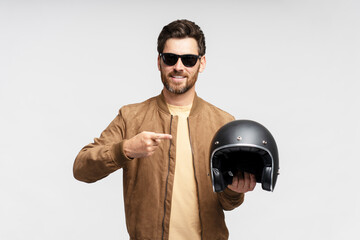 Smiling bearded biker wearing stylish sunglasses holding motorcycle helmet isolated on gray...