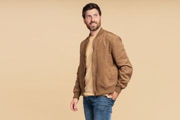 Handsome smiling bearded man wearing brown autumn jacket, stylish jeans isolated on beige background. Portrait of successful middle aged fashion model posing for pictures, studio shot