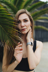 Portrait of a young girl with beautiful blue eyes and long hair dressed in sportswear, who looks at the camera and holds a palm leaf with her fingertips. - 626693627