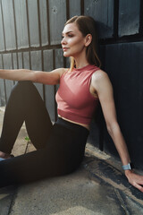 Side view of an athletic beautiful girl sitting on the floor on the street who is recovering from a morning workout. The girl runner leaned against the wall and is resting after a run.