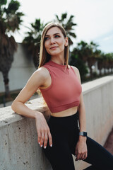 Portrait of a young smiley athletic woman in sport clothes is resting during an intense workout and stands leaning on a concrete fence while looking away.