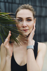 Portrait of a young attractive girl with beautiful blue eyes dressed in sportswear, wearing smartwatches, who looks at the camera and holds a palm leaf with her fingertips. - 626693674