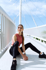 Fit and fabulous woman relaxing on the bridge after energizing online workout. A sporty attractive girl savors the sun's warmth by the bridge, marking the end of her invigorating morning jog. - 626693680