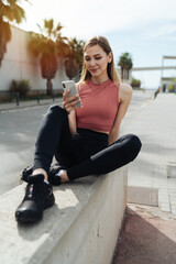 Smiling athletic woman in comfy sportswear sitting on a concrete bench while messaging on cellphone against background of palm trees.