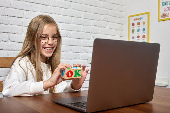 Girl in round-rimmed glasses smiles at the camera of a laptop, shows wooden cubes in her hands with colored letters. In a school classroom, a child learns English with an online teacher - Powered by Adobe