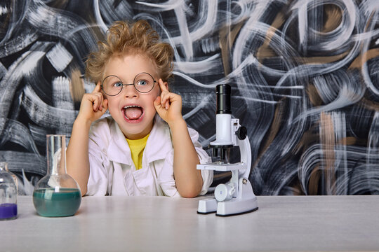 A cheerful child in glasses works with a microscope at biology lessons in a real laboratory. A boy in a bathrobe smiles happily while sitting at a table on which stands a microscope on a white tripod