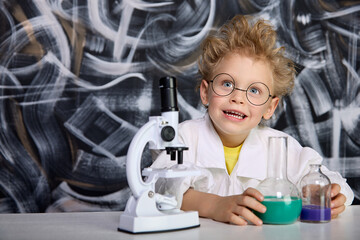 An enthusiastic Caucasian child is sitting at a table with test tubes and a microscope. Boy with...