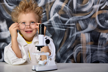 Boy holds in his hands glasses scientist in children's laboratory with real tools for conducting research. Young scientist poses for camera against background chalk board and with microscope on table