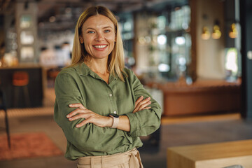 Stylish middle-aged businesswoman standing with crossed hands on coworking space background