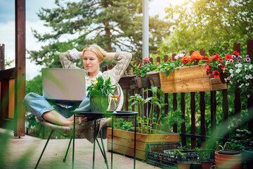 Fototapeta na wymiar A beautiful middle-aged blonde woman in her country house is sitting on the terrace surrounded by flowers and working on a laptop. The life of downshifter in a house in nature surrounded by flowers