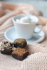 Delicious pieces of chocolate brownies on soft knitted fabric with a blurry cup of marshmallow on the background. Selective focus