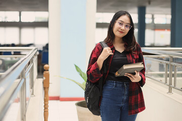 Pretty smart young Asian student standing confidently and smiling to the camera, hold book, against library background