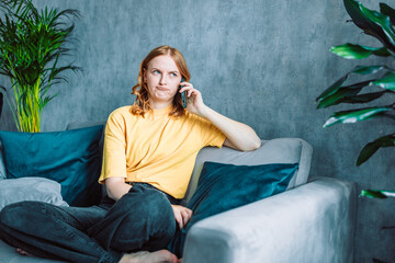 Unhappy worried woman talking on phone while sitting on sofa at home. Sad 30s woman has difficult phone talk hold gadget by ear listen to unexpected bad news think on answer.