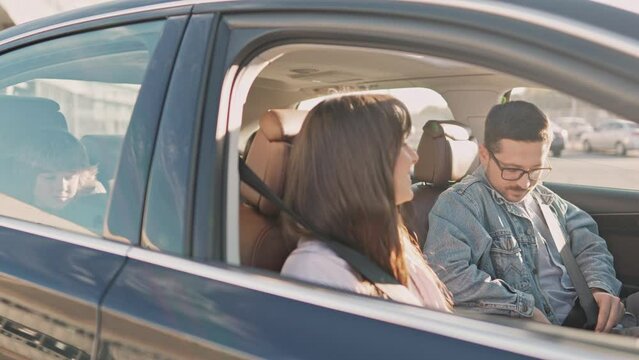 Cheerful young family is preparing for family trip by car. Caring parents fasten their seat belts. Caring for safety while traveling on the road. Concept of tourism and road trip.