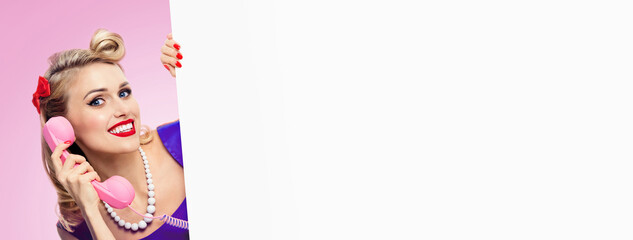 Image of woman with phone, in pin up style dress, showing blank signboard with copyspace area, over pink background. Female blond model posing in retro fashion and vintage studio concept.