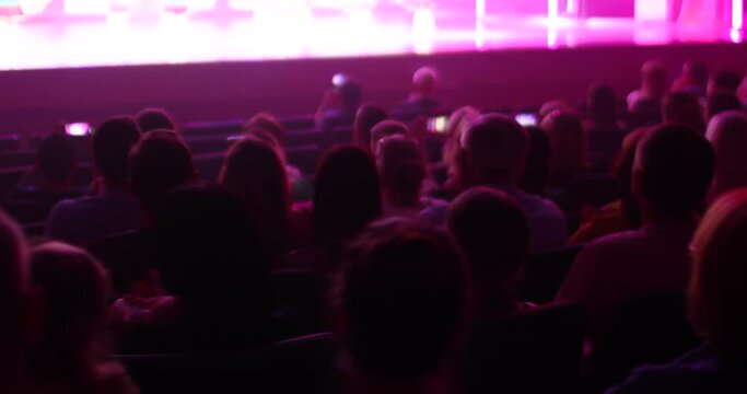 Back view of people sitting in a full hall and applauding. Indoors. Spectators watch the performance in subdued purple light. The concept of holding events, concerts, holidays.