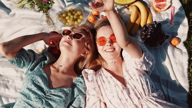 Two young beautiful smiling hipster female in trendy summer sundress and hats. Carefree women making picnic outside. Positive models sitting on plaid on grass, hugging, eating fruits. Top view