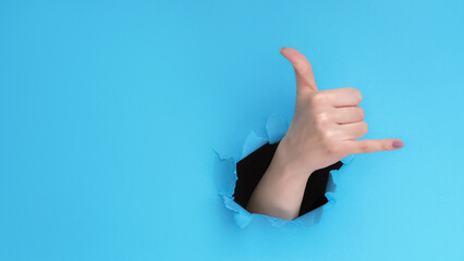 Call gesture. Shaka sign. Hang loose. Female hand showing greeting signal inside breakthrough hole on blue torn paper wall background with empty space.