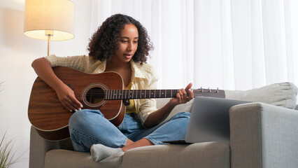 Online school. Guitar lesson. Remote learning. Woman guitarist practicing string instrument internet class at laptop sitting couch at home room.