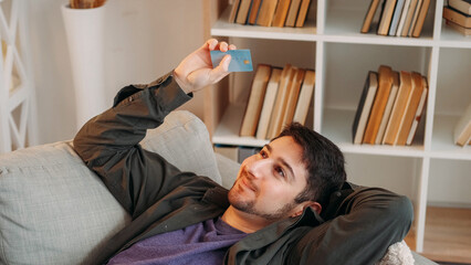 Credit card service. Bank customer. Plastic debit. Joyful man relaxing on sofa satisfied with buying online purchase cash back bonus financial loan at home.