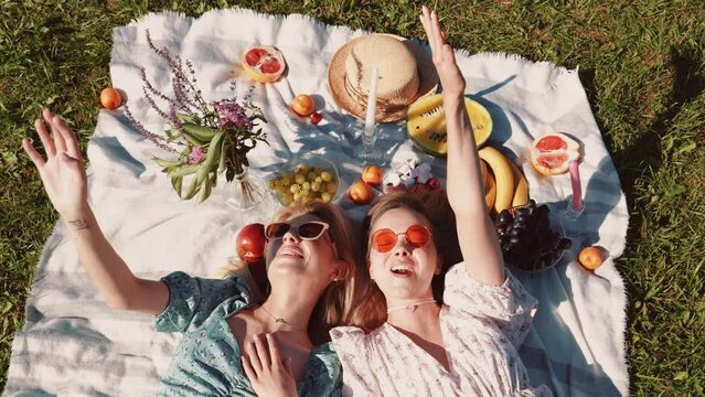 Two young beautiful smiling hipster female in trendy summer sundress and hats. Carefree women making picnic outside. Positive models sitting on plaid on grass, hugging, eating fruits. Top view