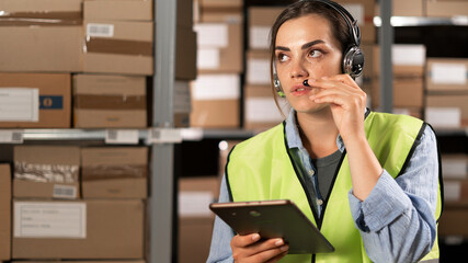 brazilian female worker in headset at logistics warehouse using digital tablet. speaks into a microphone against the background of shelves with cardboard boxes.
