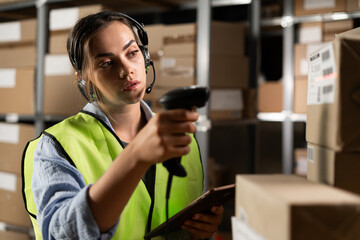 brazilian woman worker in a headset in a logistics warehouse using a digital tablet looking for the necessary product. against the background of shelves with cardboard boxes scans a barcode