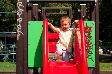 Little toddler playing on the playground and sitting on a slide - obrazy, fototapety, plakaty