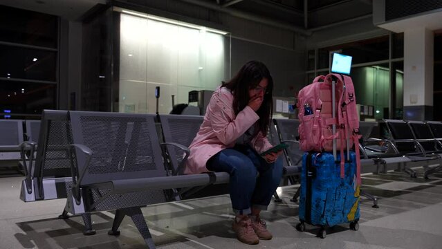 Woman waits her flight next to luggage playing with phone while coughs. Pan left