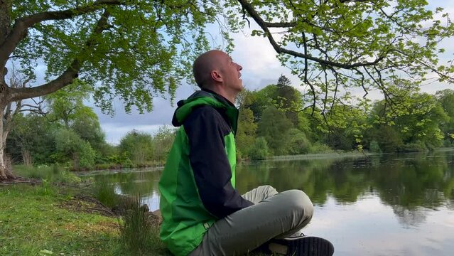 Bald man breathwork near lake. Hand on knees under tree. Wim Hof method breathing practise around nature inner work. Full side body undressing green jacket. Closeup. Black jumper
