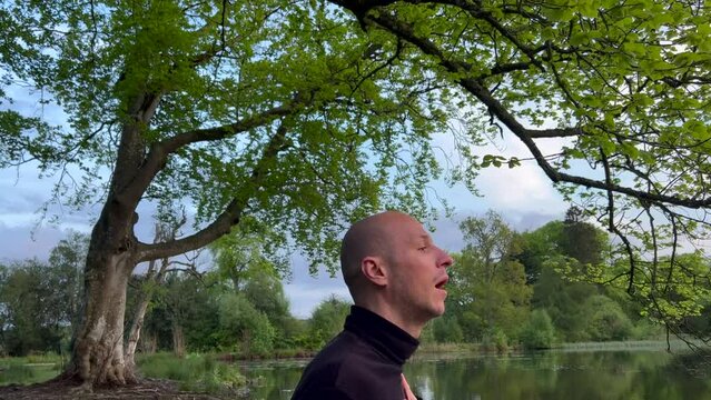 Young bald man breathwork near lake. Hand on heart under tree. Wim Hof inner work method breathing practise around nature. Dressed with turtleneck black jumper. Pan up shot exploring environment