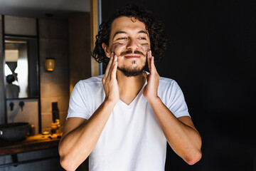 latin man applying exfoliating facial mask treatment and taking care of skin in loft apartment in...
