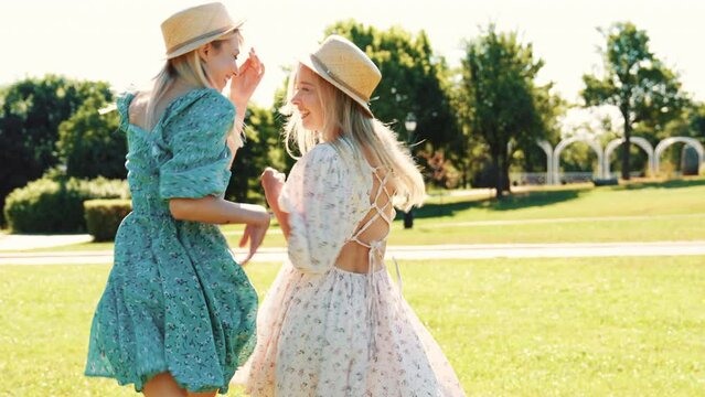 Two young beautiful smiling hipster woman in trendy summer sundress. Carefree women posing in the park in hats. Positive models spinning around and run away. Following camera